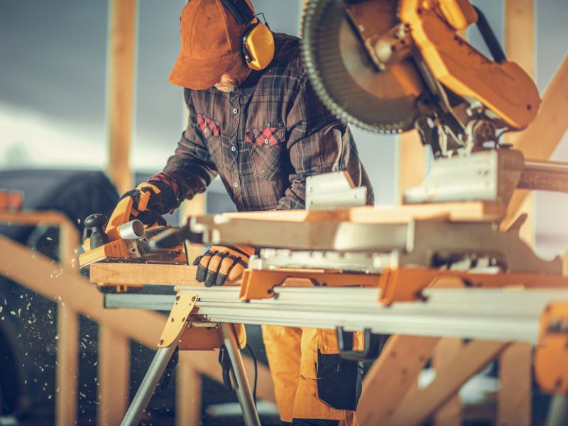 Caucasian Construction Worker in His 40s with Noise Reduction Headphones on His Head Planing Wood with Electric Planer Inside Newly Built Wooden Skeleton Frame of House.